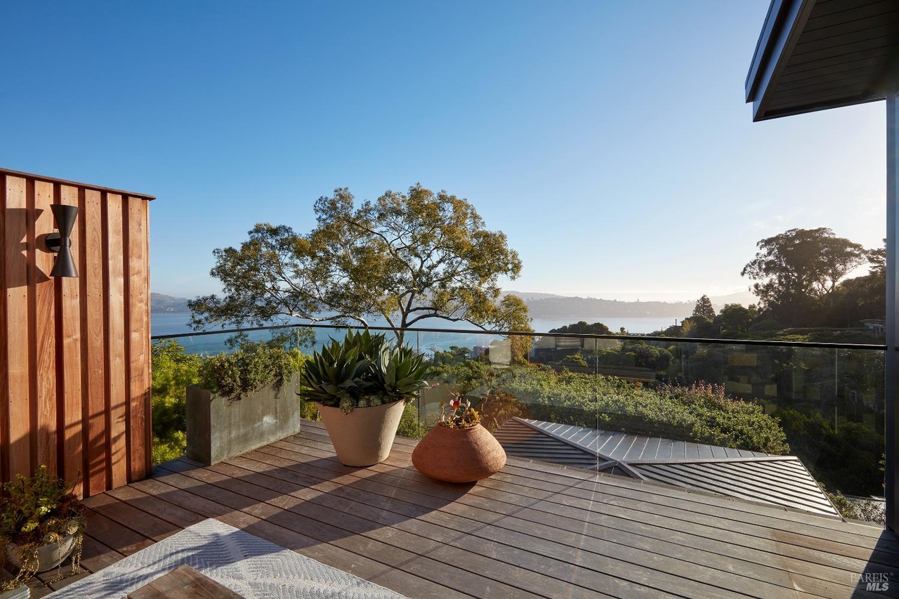 A wood-decked terrace with a glass railing, a sculptural agave in a stone planter, and a specimen tree framing Richardson Bay in soft afternoon light