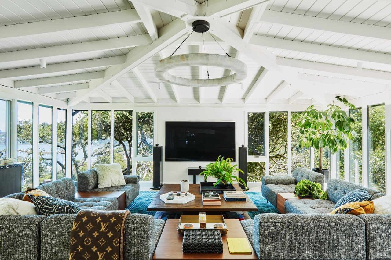 A family room under a vaulted white-painted beamed ceiling, a circular sculptural chandelier above gray sofas, walls of glass on three sides opening to the oak canopy