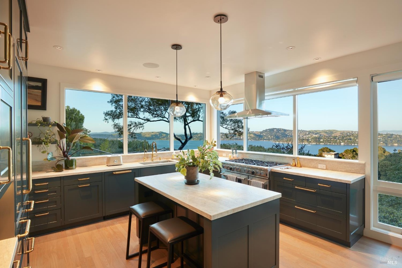 A kitchen with deep teal cabinetry and brass hardware, a marble island and stainless range, wraparound windows framing Richardson Bay through the trees at dusk