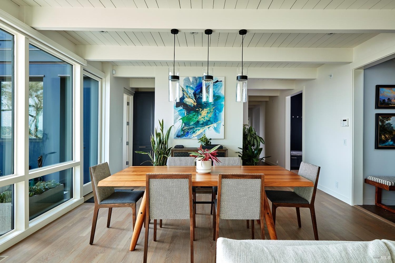 A warm wood dining table with woven chairs beneath three glass pendant lights, a blue and green abstract painting on the wall, exposed beams overhead