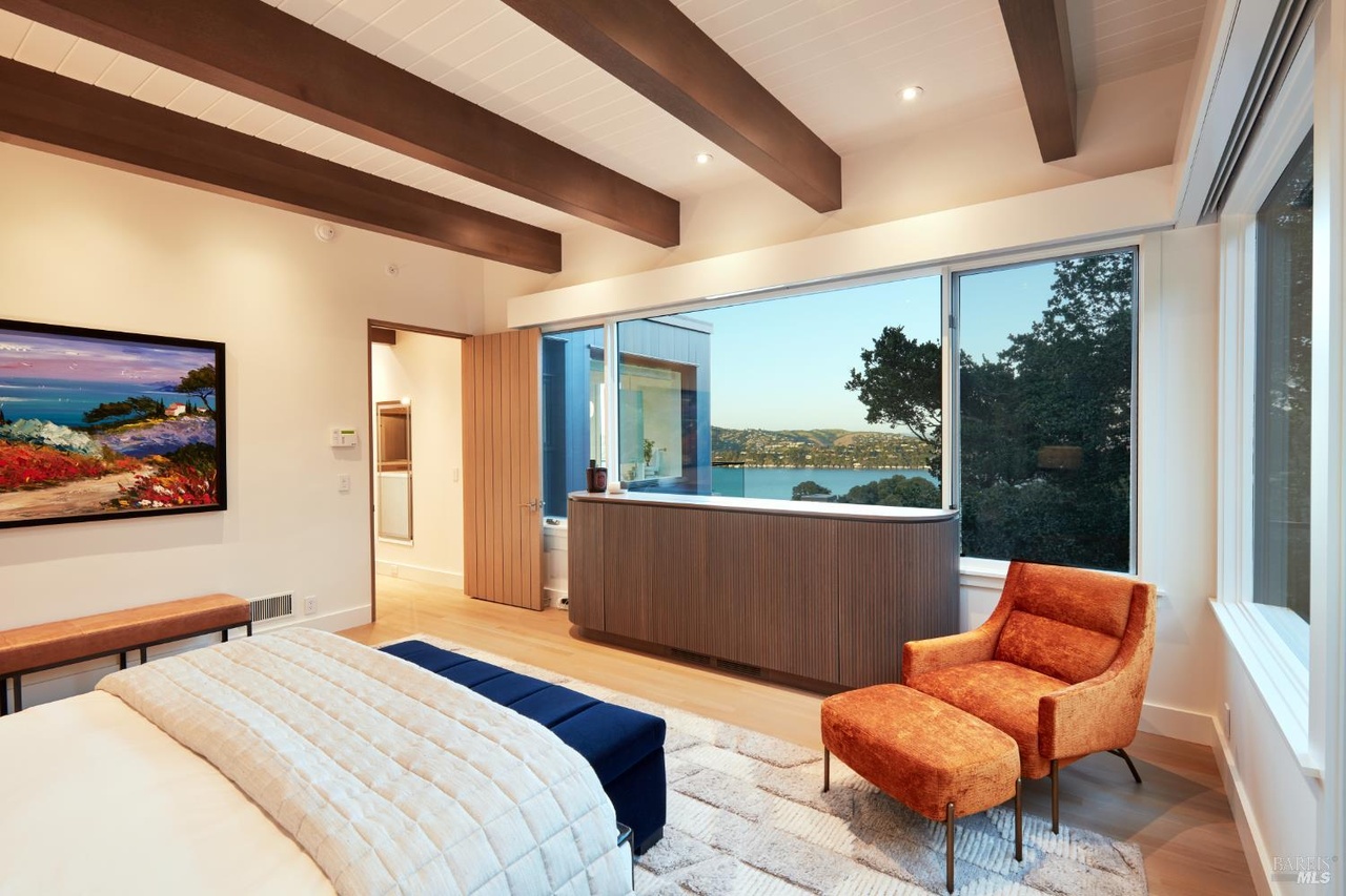 The primary bedroom at dusk with an orange velvet lounge chair and ottoman, exposed wood beams, a curved wood-slat partition, and corner walls of glass over the bay