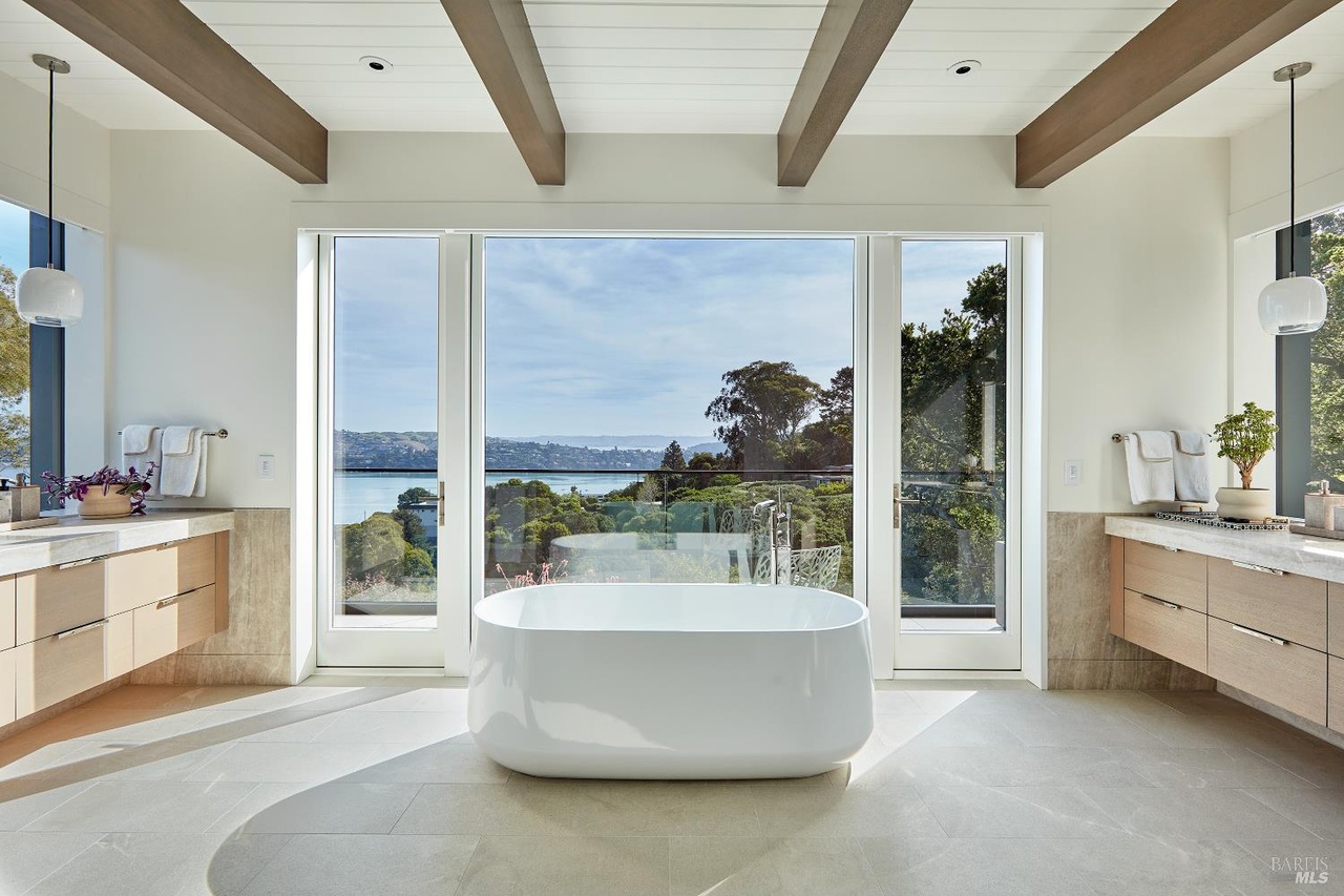 A spa-inspired bath with a freestanding white soaking tub centered on floor-to-ceiling glass over the bay, exposed wood beams, and twin floating oak vanities on either side