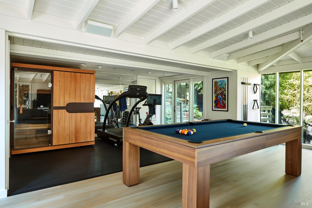 A lower-level wellness and games room with a wood-frame billiards table in navy felt, a treadmill and exercise bike, an infrared sauna cabinet, and glass walls to the greenery