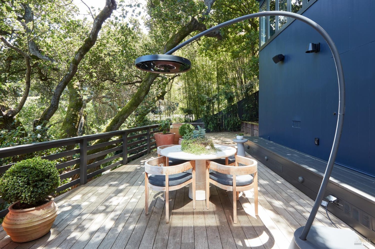 A garden dining deck with a round cafe table and wood-and-cushion chairs, a sculptural arc patio heater, set against the deep blue facade and screened by oak and bamboo